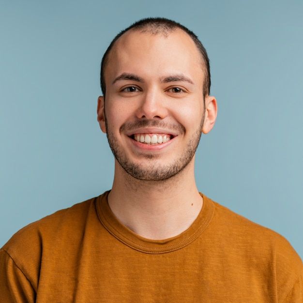 http://Portrait%20of%20a%20happy%20young%20man%20with%20a%20shaved%20head%20and%20trimmed%20beard,%20wearing%20a%20mustard-colored%20shirt%20and%20smiling%20confidently%20against%20a%20blue%20background.